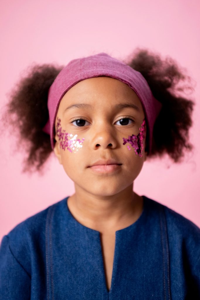 A young girl with glitter stickers on cheeks, posing against a pink background.